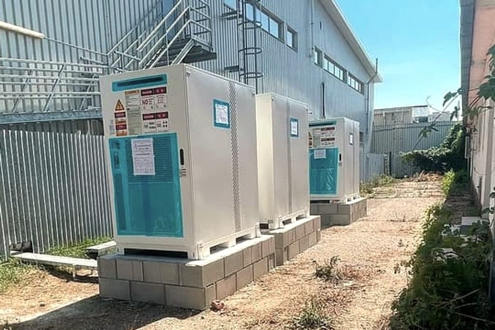 Drei Batterieschränke eines Speichers auf Betonsockeln neben einer Halle